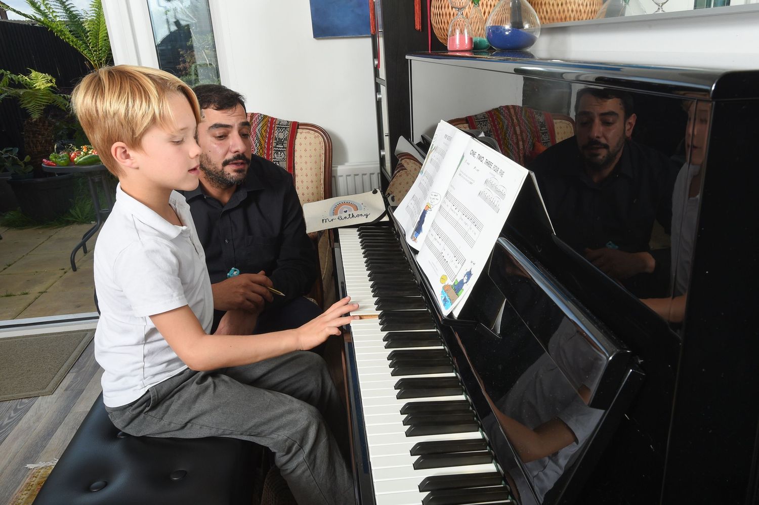 A young boy plays the piano, focused on sheet music, while an adult sits beside him giving instructions.