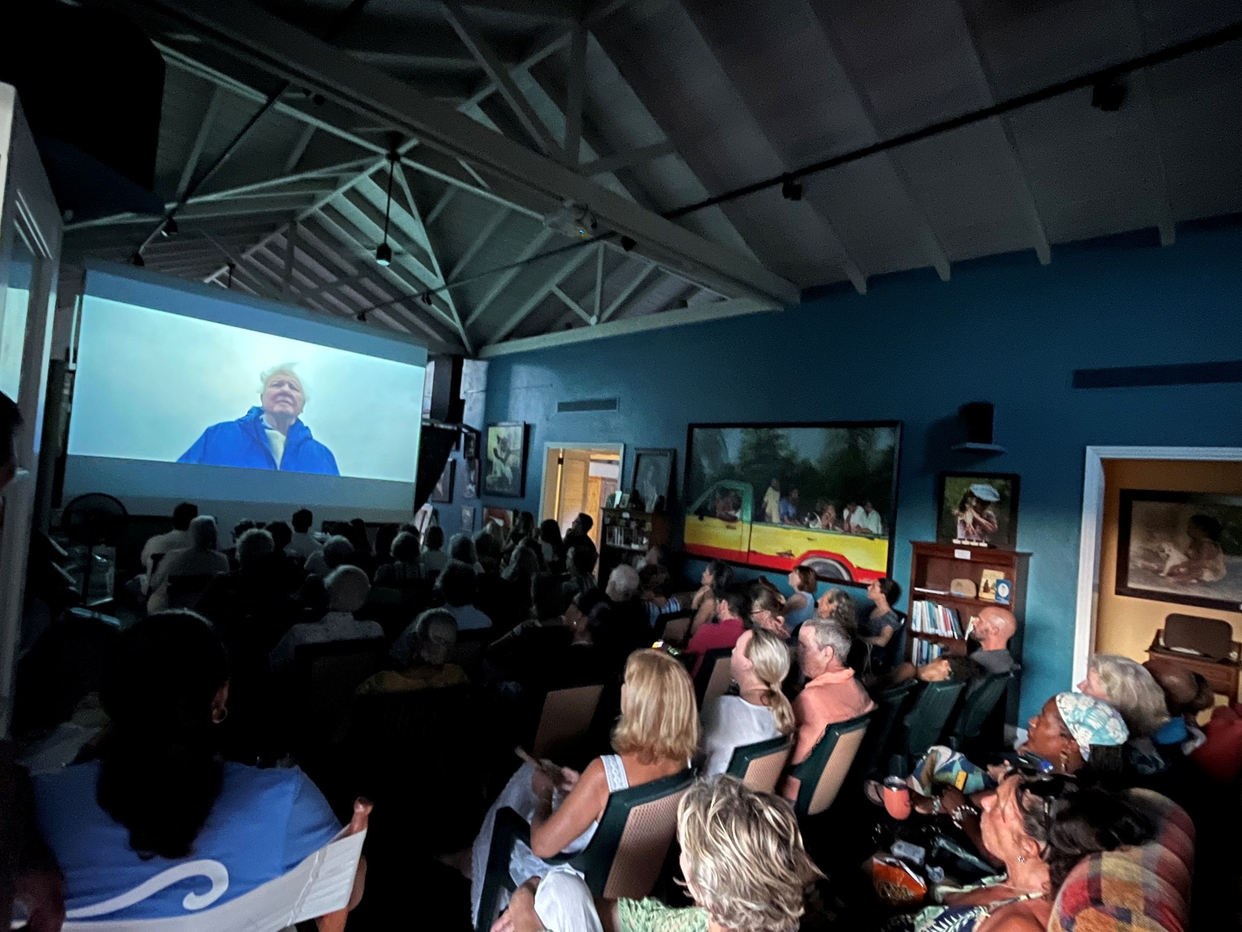 Crowded indoor event with audience watching a video presentation on a large projection screen in a blue-walled venue.