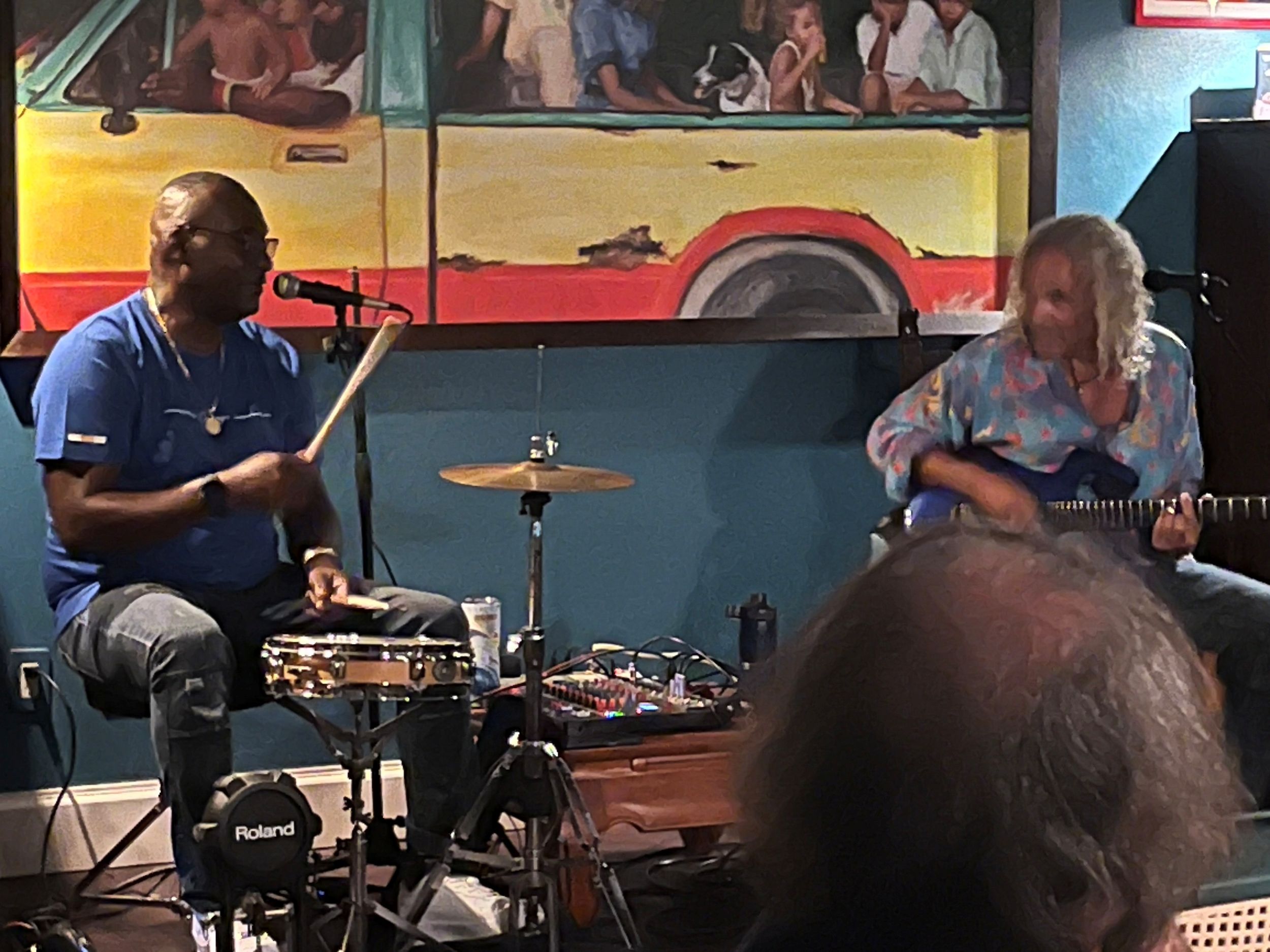 Two musicians perform live, one playing drums and another on guitar, in front of a colorful painted bus mural.