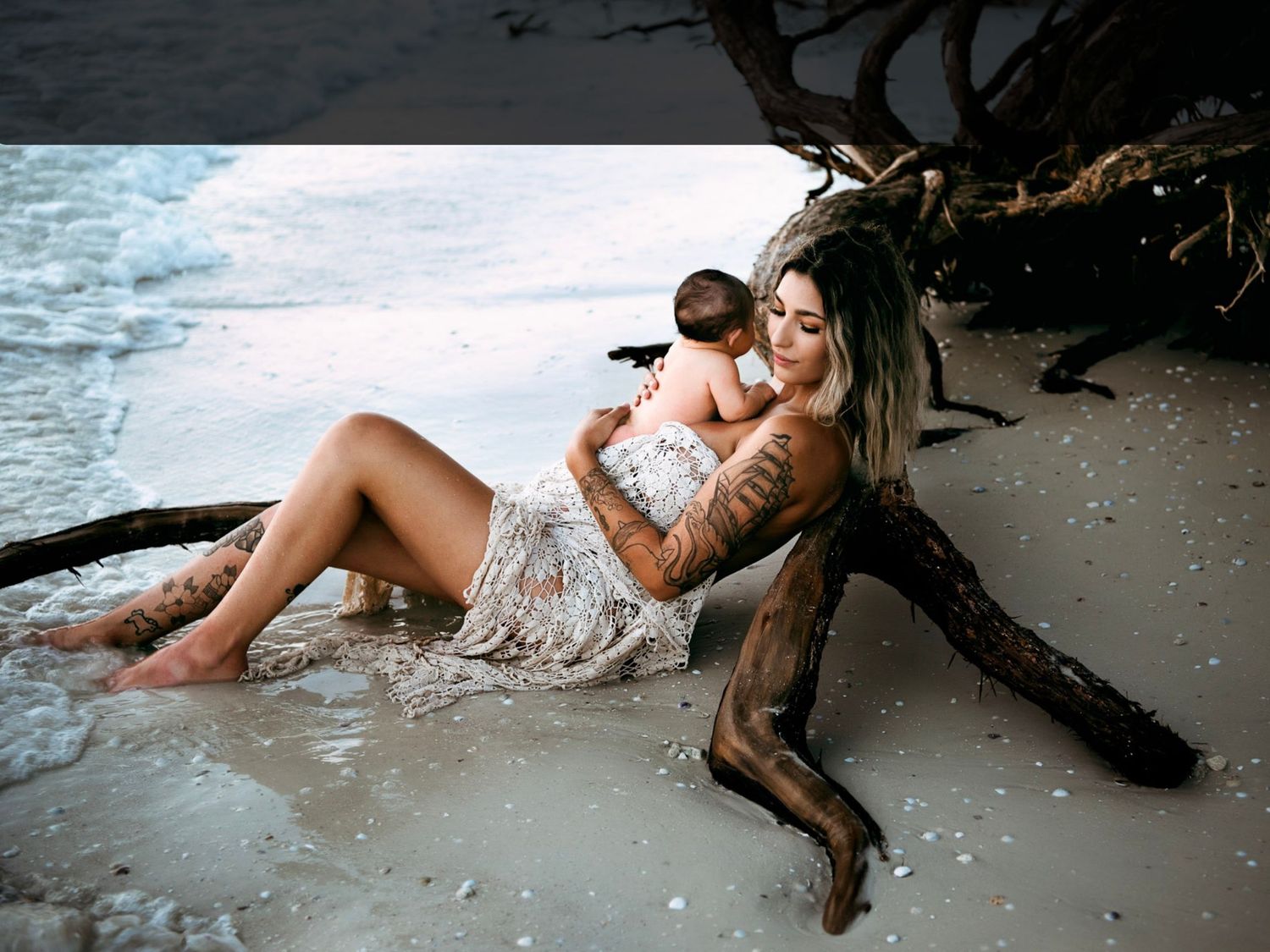 Tattooed mother cradling newborn baby on sandy beach beside driftwood with ocean waves in background.