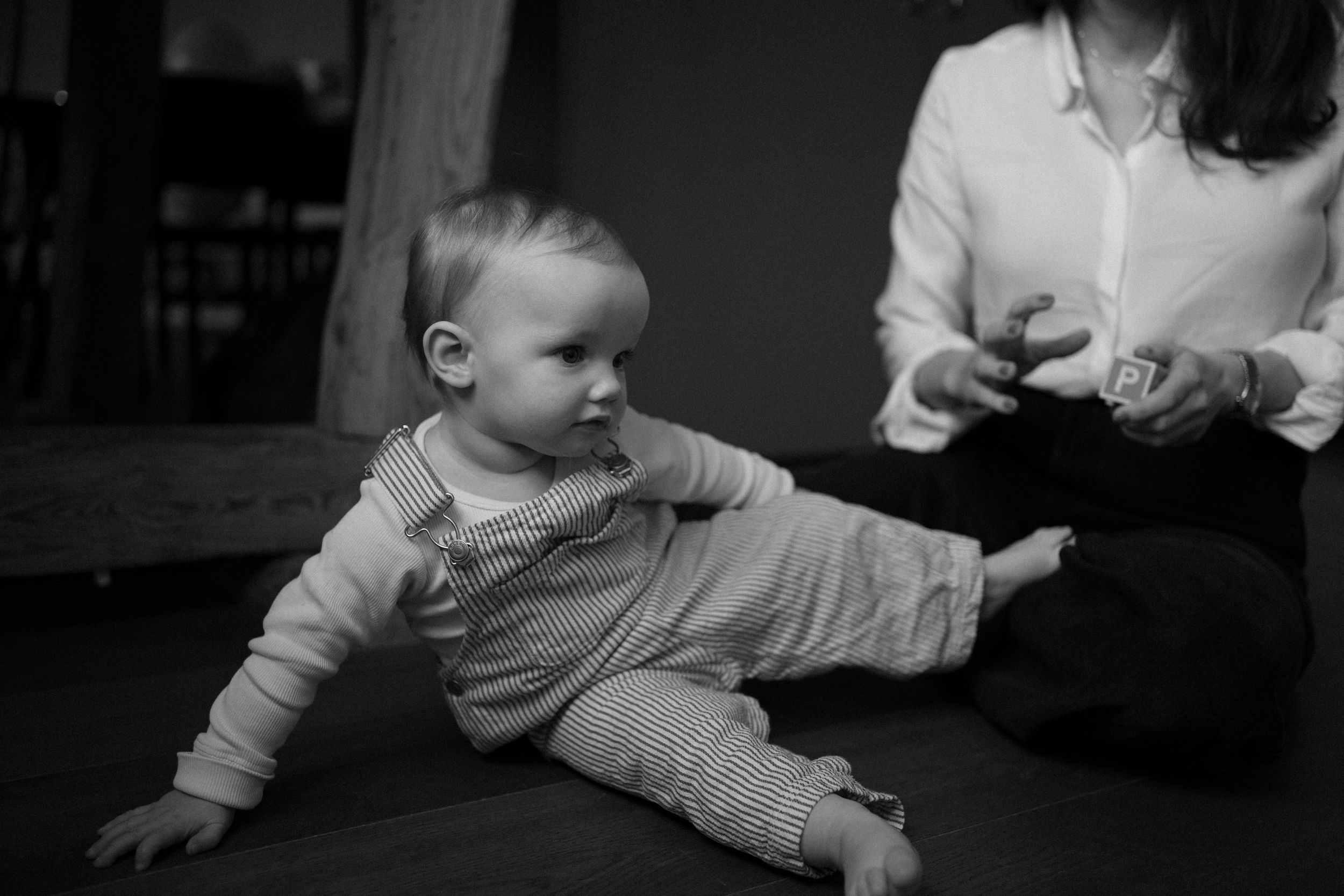 Ett litet barn sitter på golvet med hängselbyxor. Bakom sitter hennes mamma med en träkloss med bokstaven P.
