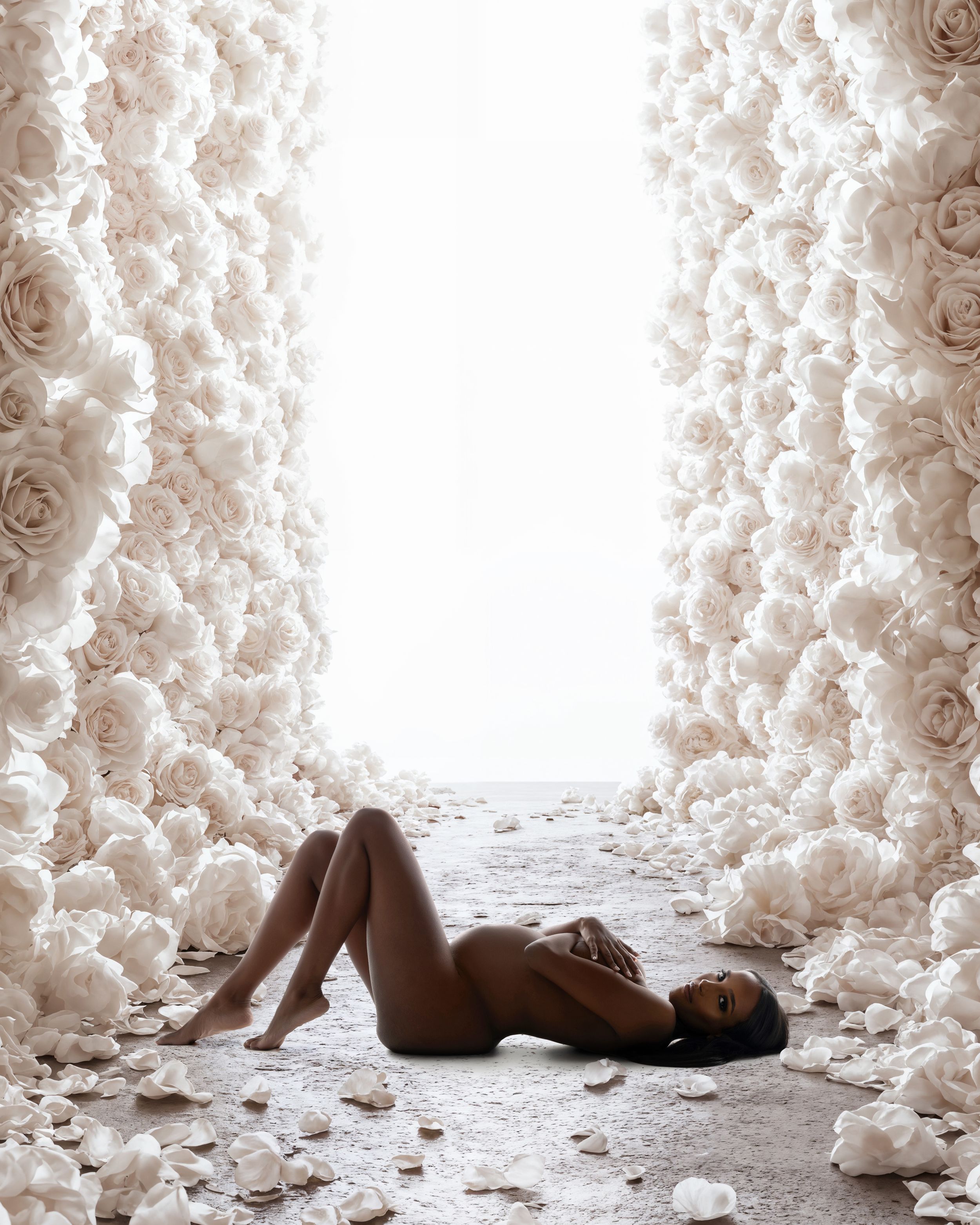Artistic photo of a nude figure laying on the ground surrounded by white roses forming a tunnel-like pathway.
