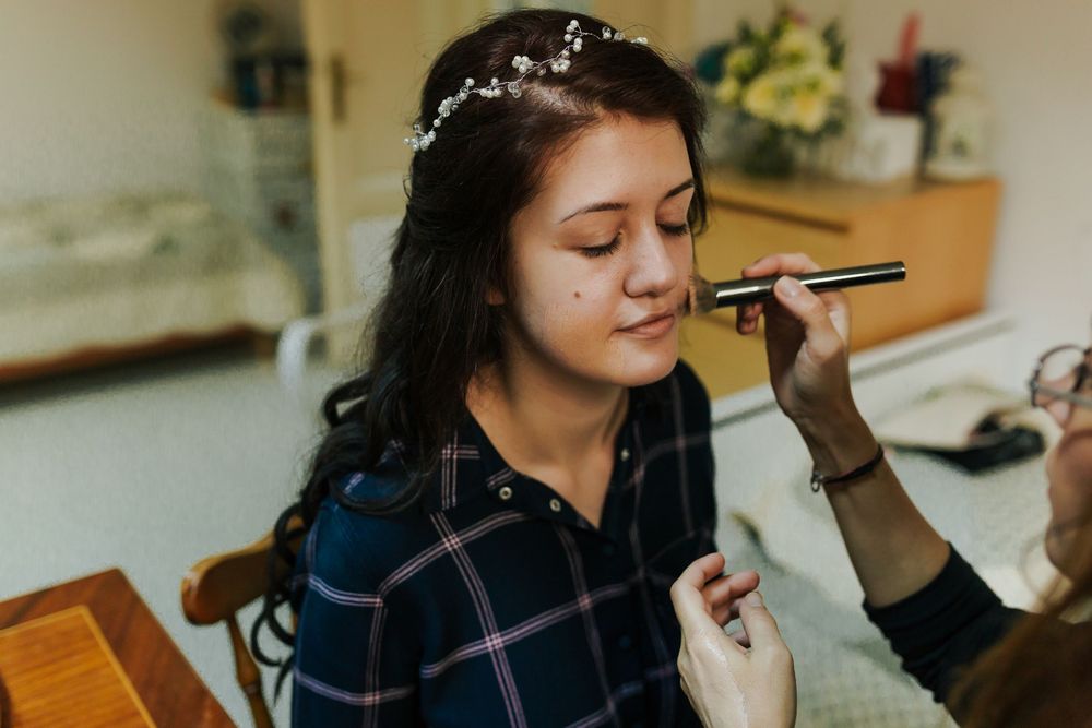 Makeup artist applying cosmetics to young brunette bride with floral hair accessory during wedding preparation.