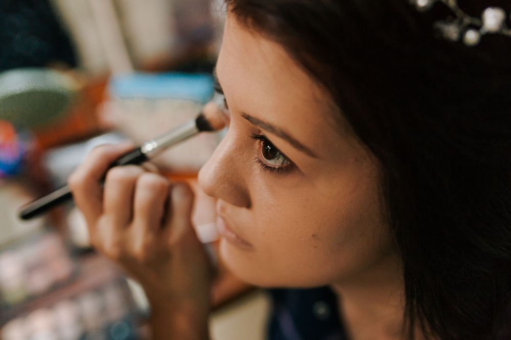 Close-up of makeup artist applying eyeshadow to brunette bride with floral hair accessory before wedding.