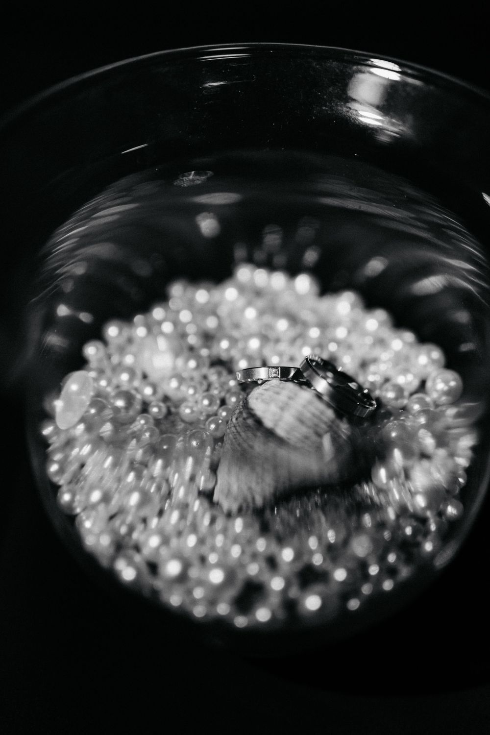 Black and white artistic photo of wedding rings resting on pearl beads inside a glass bowl.