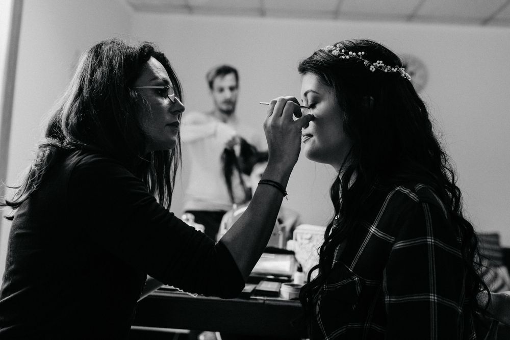 Black and white photo of makeup artist applying eyeshadow to bride wearing floral crown before wedding.