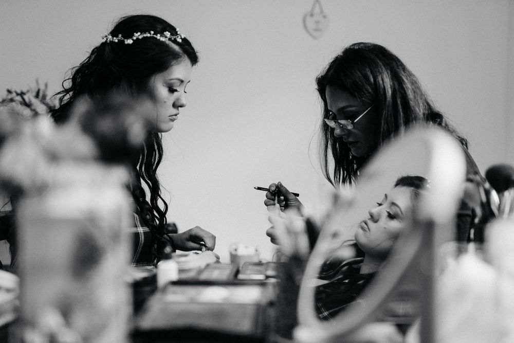Black and white photo of bride in floral crown getting makeup done reflected in vanity mirror.