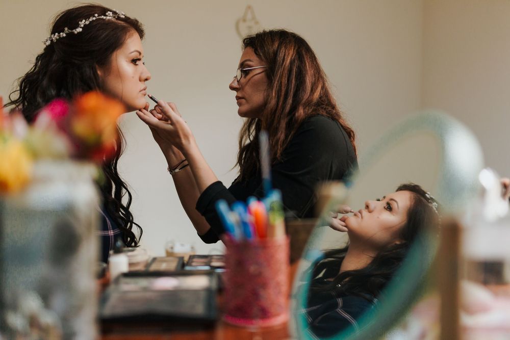 Makeup artist applying foundation to bride wearing floral crown while assistant watches in mirror.