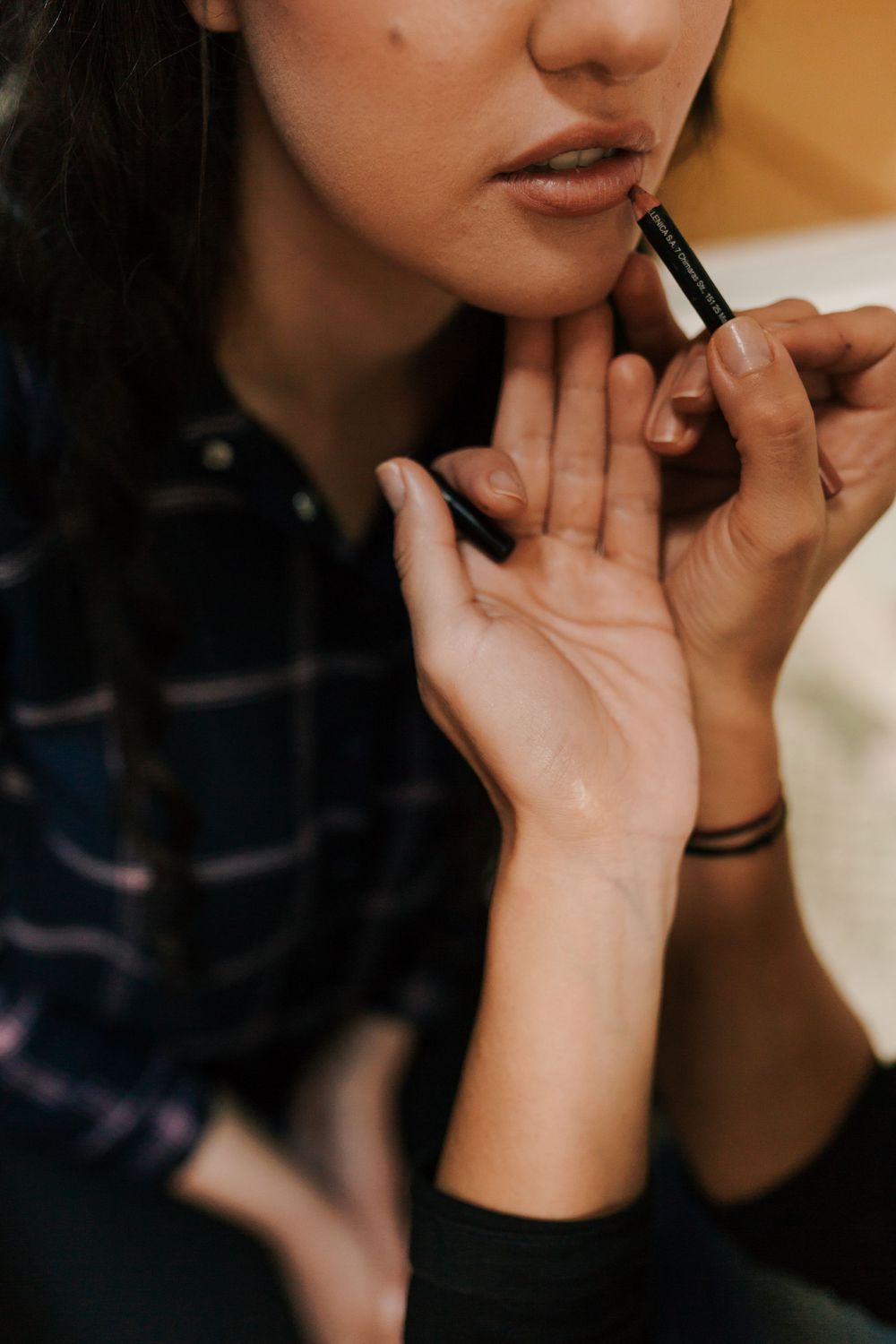 Close-up of makeup artist carefully applying lip liner to bride in plaid shirt before wedding.