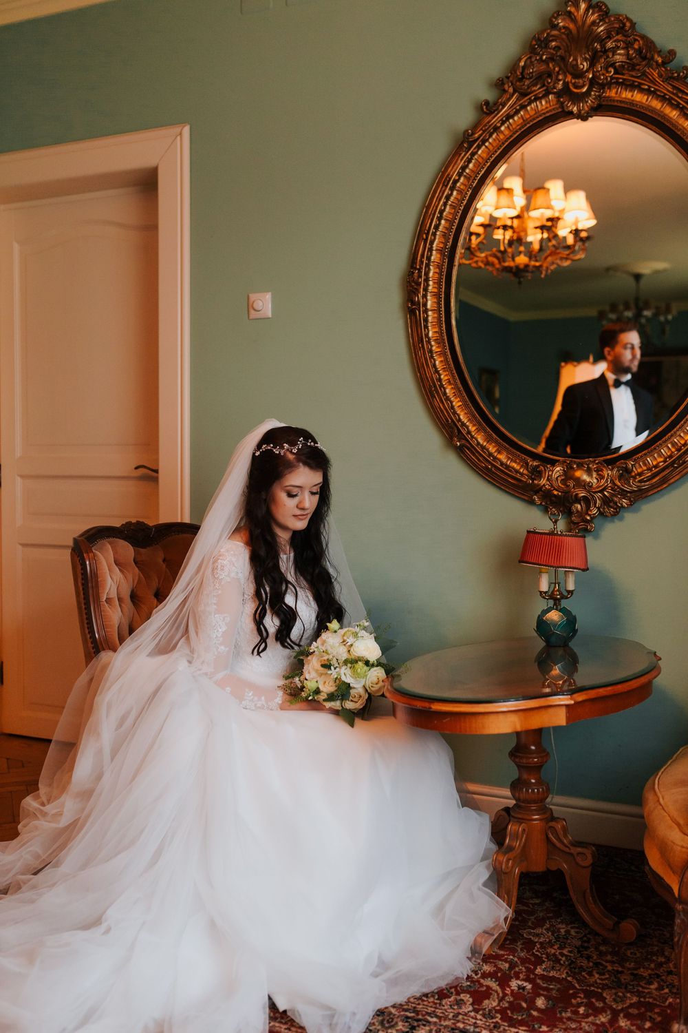Bride in white gown sits by ornate gold mirror, holding bouquet, groom reflected in background.