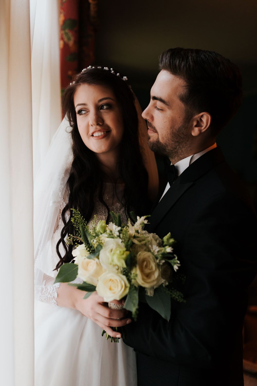 Bride and groom embrace near window, groom in tuxedo holding white rose bouquet, bride in veil.
