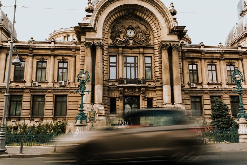 Blurred car passes in front of grand historic neoclassical building facade with ornate architectural details.