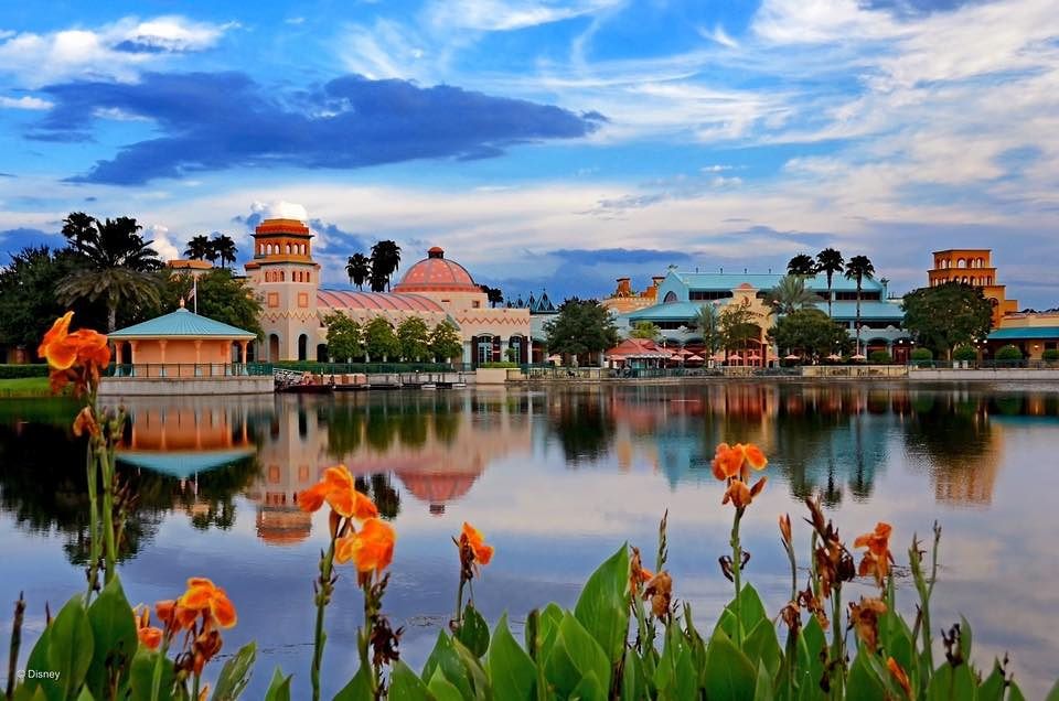Scenic lakeside view of a resort with Spanish-style architecture reflecting in calm waters at sunset.