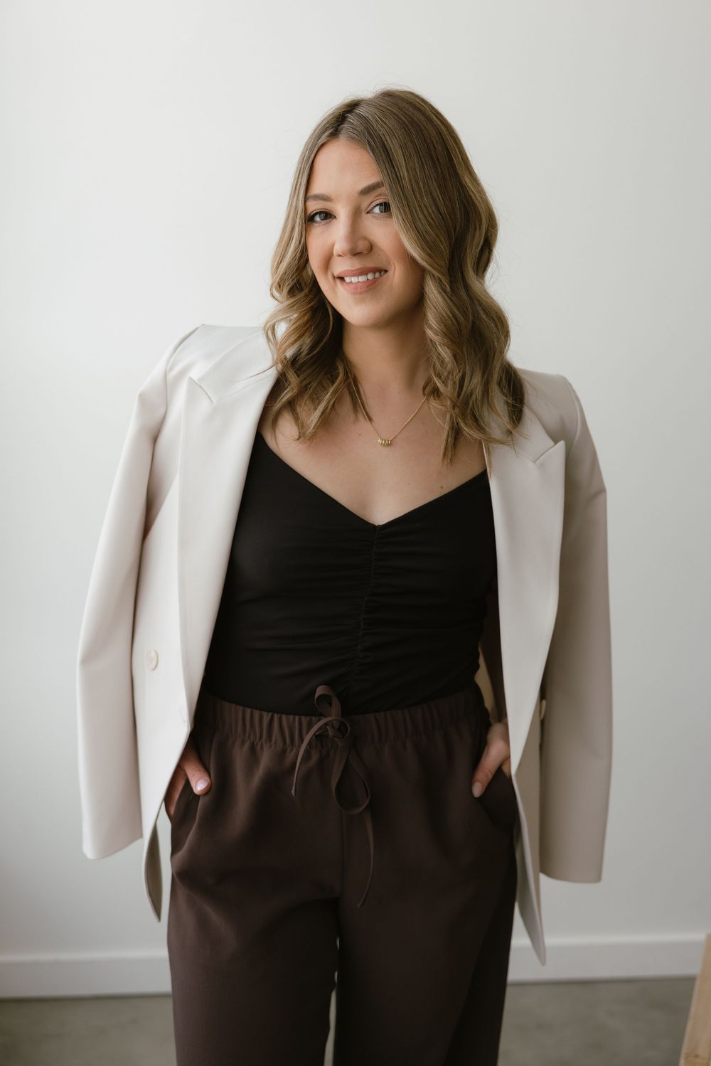 Smiling woman in black top, cream blazer, and brown drawstring pants poses confidently with hands in pockets.