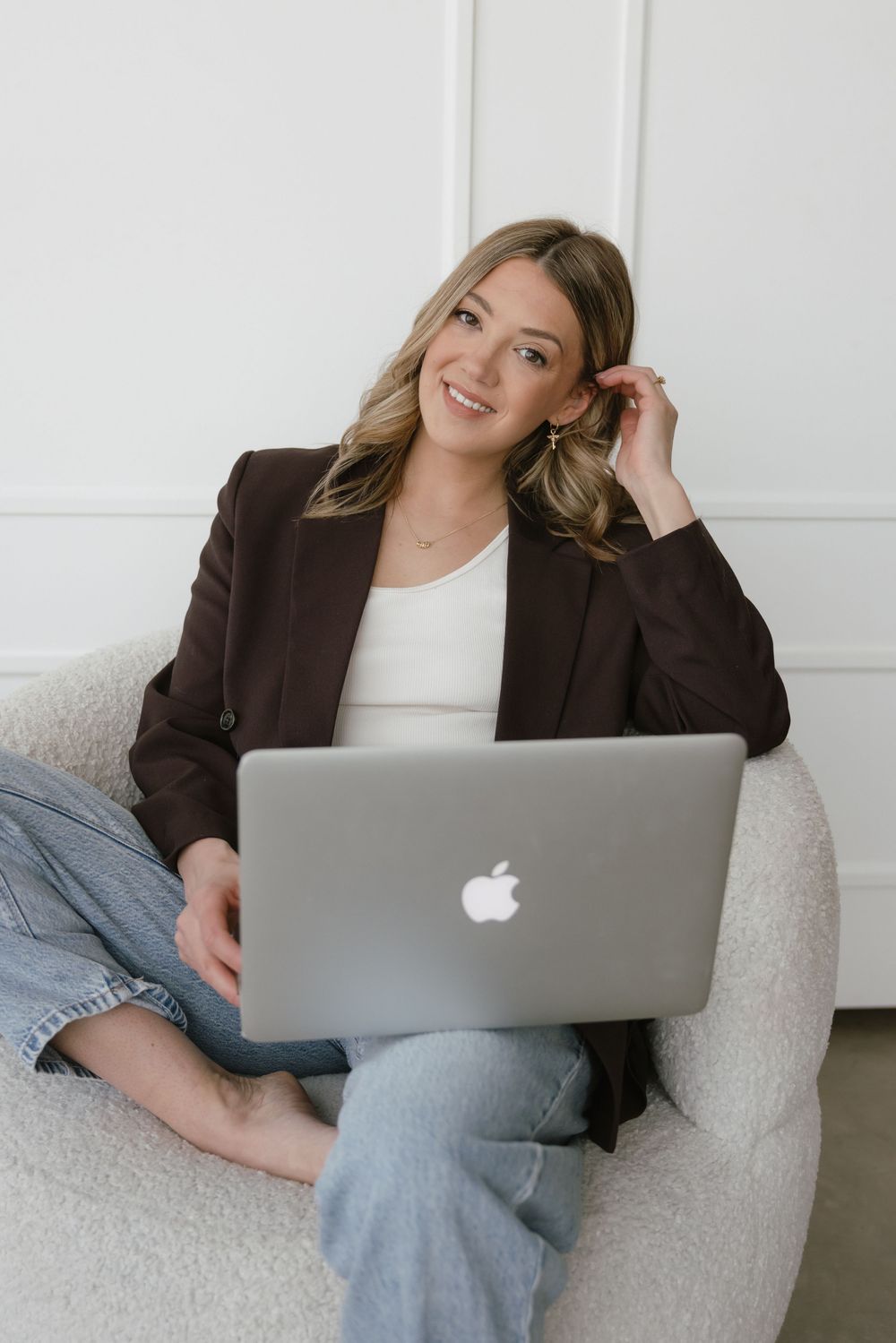Woman in brown blazer smiling with MacBook laptop while sitting in white chair.
