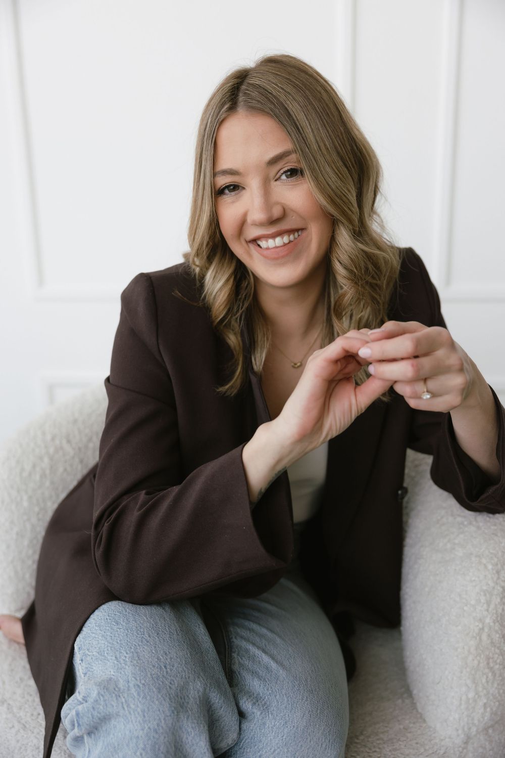 Smiling woman in dark brown blazer and jeans sitting in white chair with hands clasped in bright studio setting.