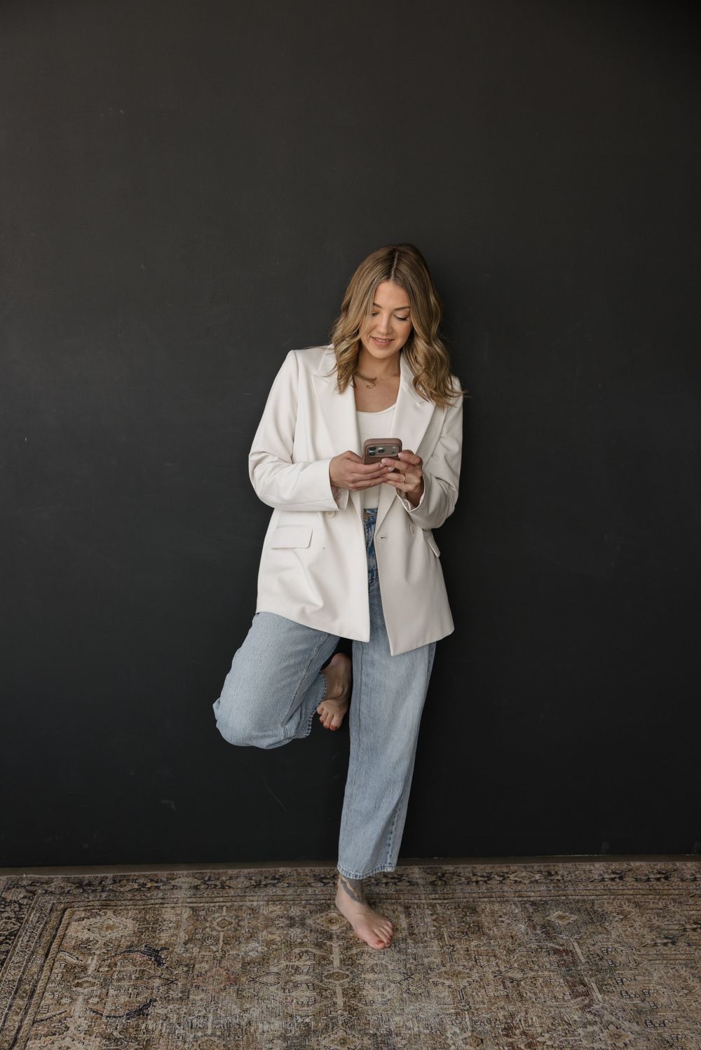 Woman in white blazer and jeans standing on one foot while using smartphone against dark wall.