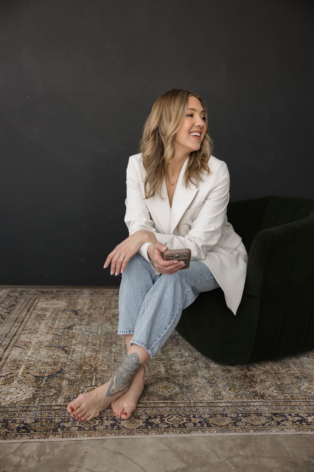 Smiling woman in white blazer and jeans sitting on floor holding smartphone near green ottoman.