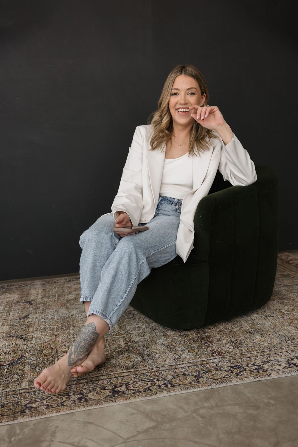 Smiling woman in white blazer and jeans sits barefoot on dark chair, holding phone, with tattoo on ankle.