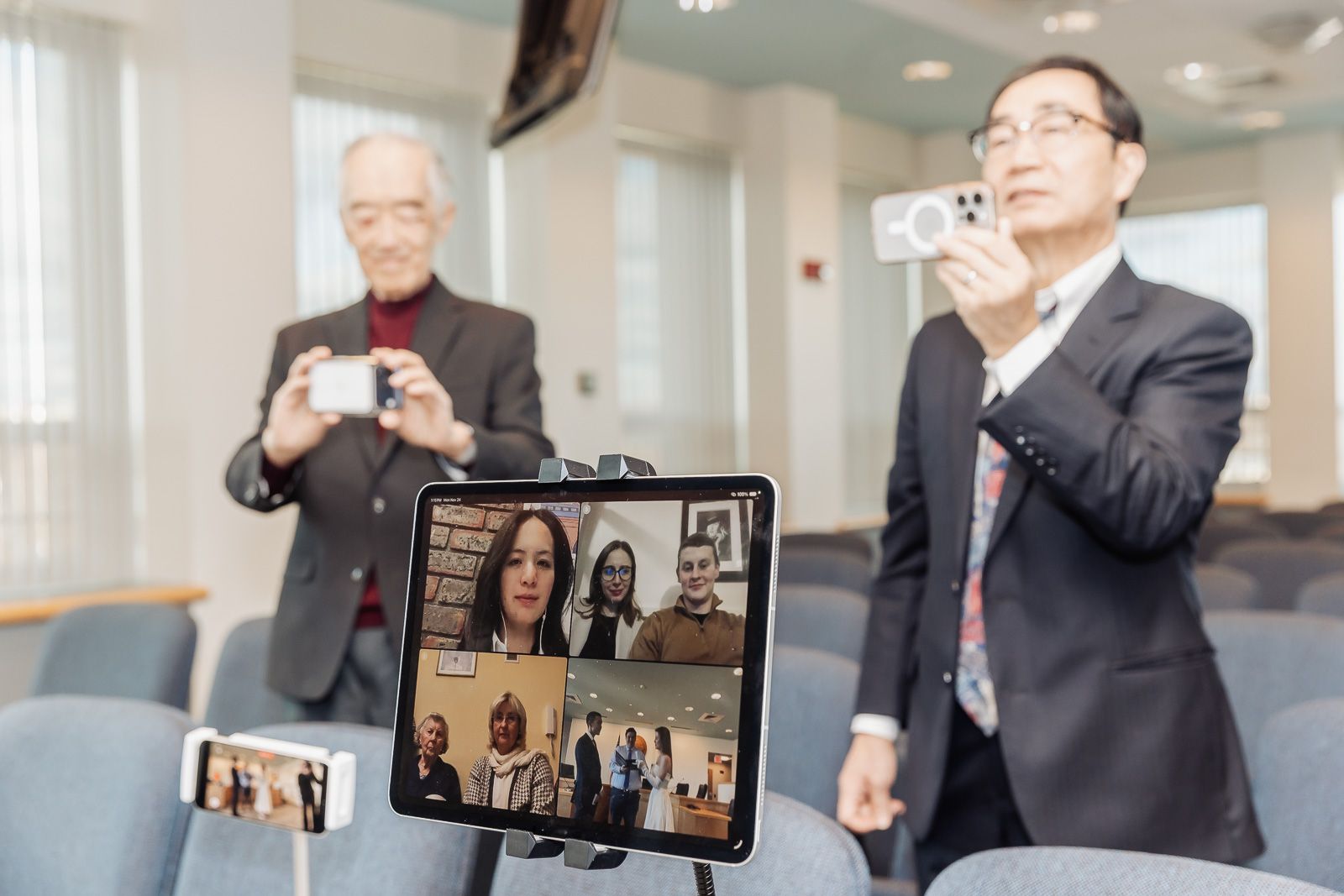 Two men photograph a tablet displaying a video call with multiple participants in a modern conference room.