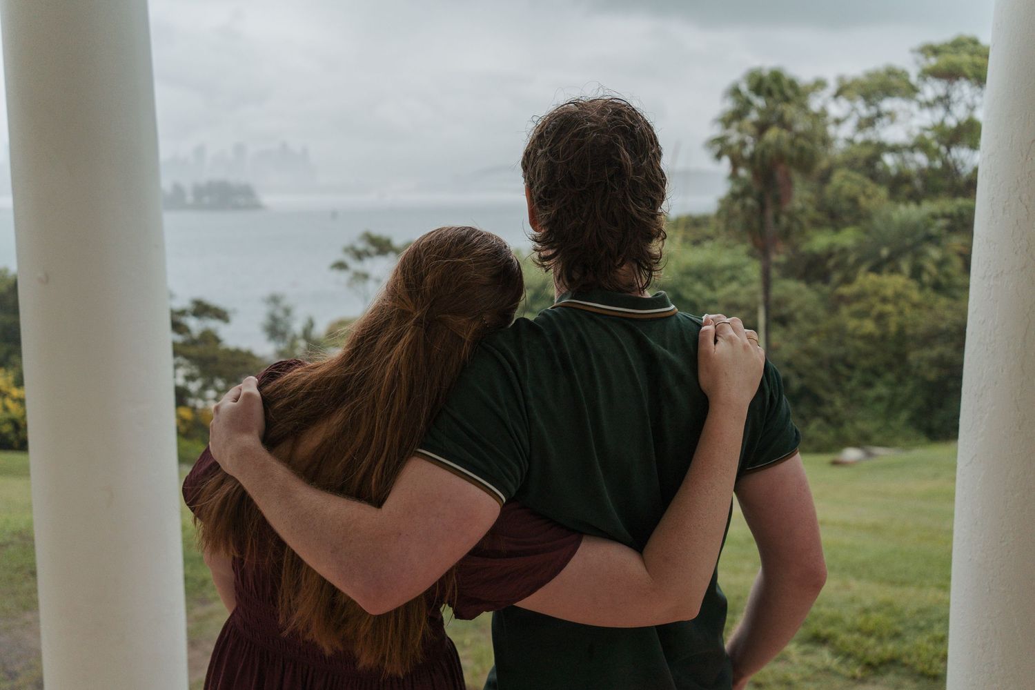 A couple embraces while gazing at a scenic ocean view from a white-columned porch surrounded by lush greenery during engagement photoshoot