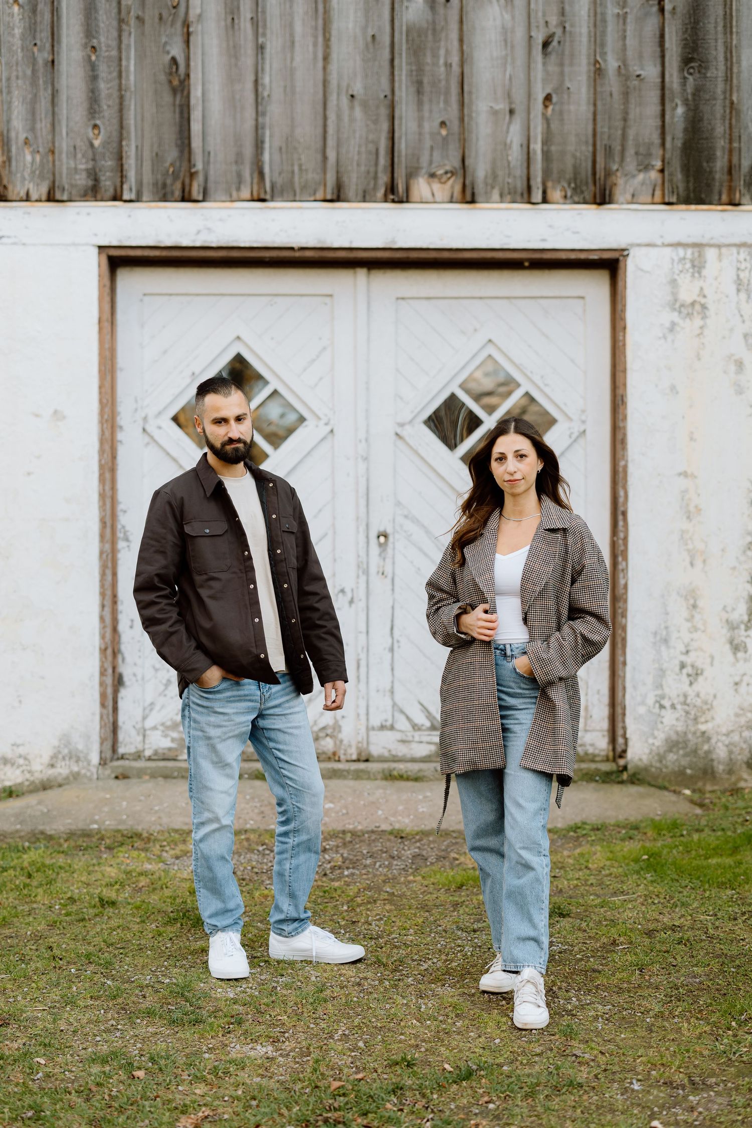A bearded man and a woman in casual jackets and jeans stand before a rustic white barn door.