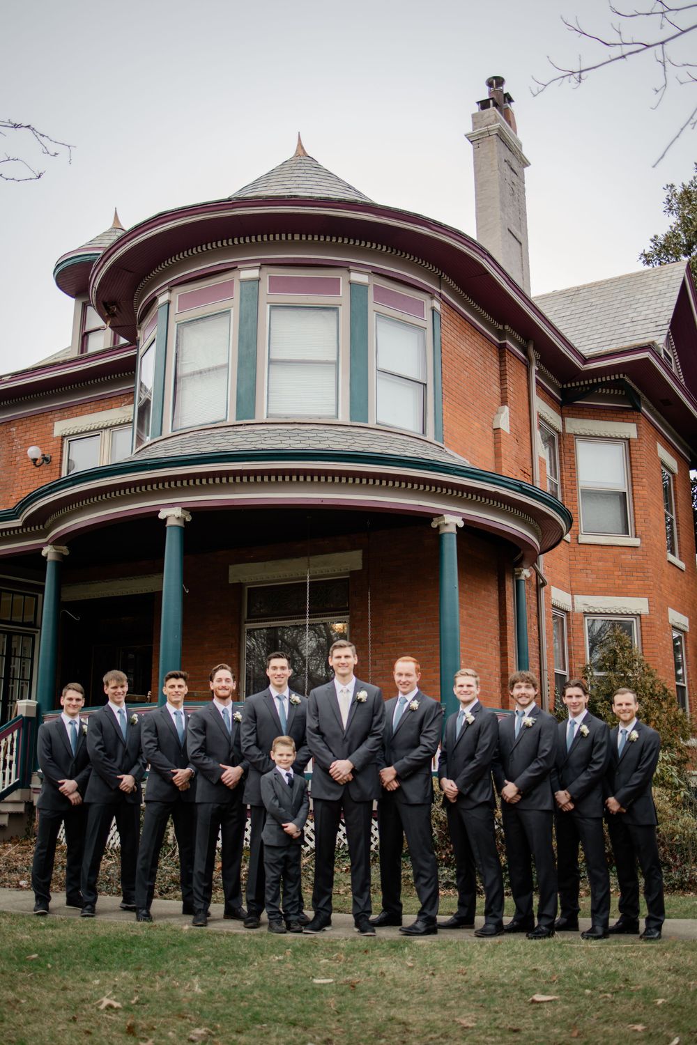 Groomsmen in dark suits pose outside a Victorian brick mansion with a rounded turret and teal columns.