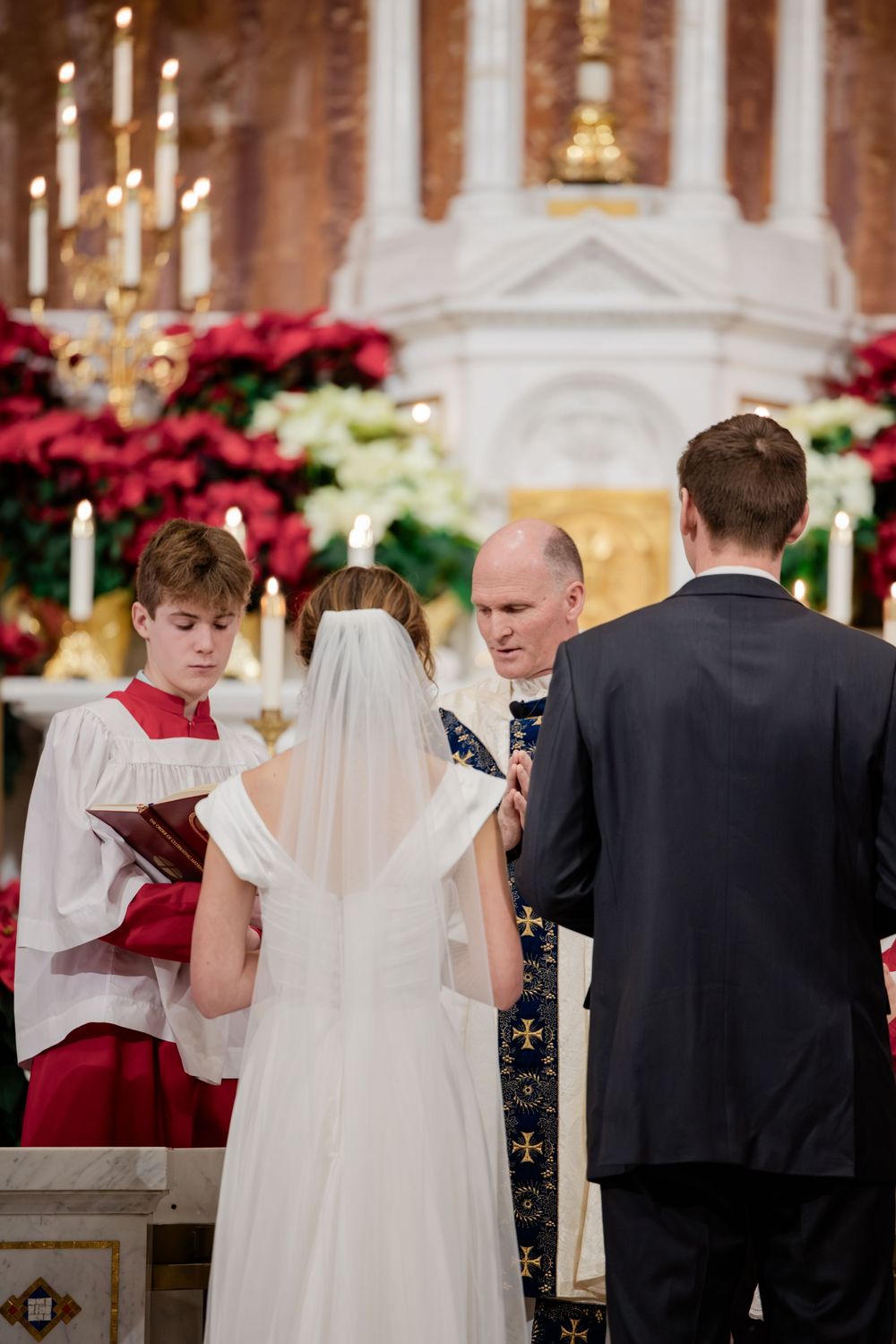 Bride and groom exchanging vows at a church altar with an officiant and altar server, surrounded by poinsettias and candles.