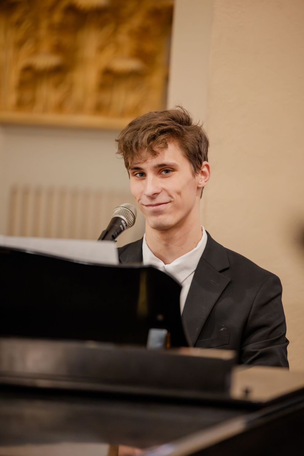 Young male pianist in black suit smiling at microphone during indoor performance.