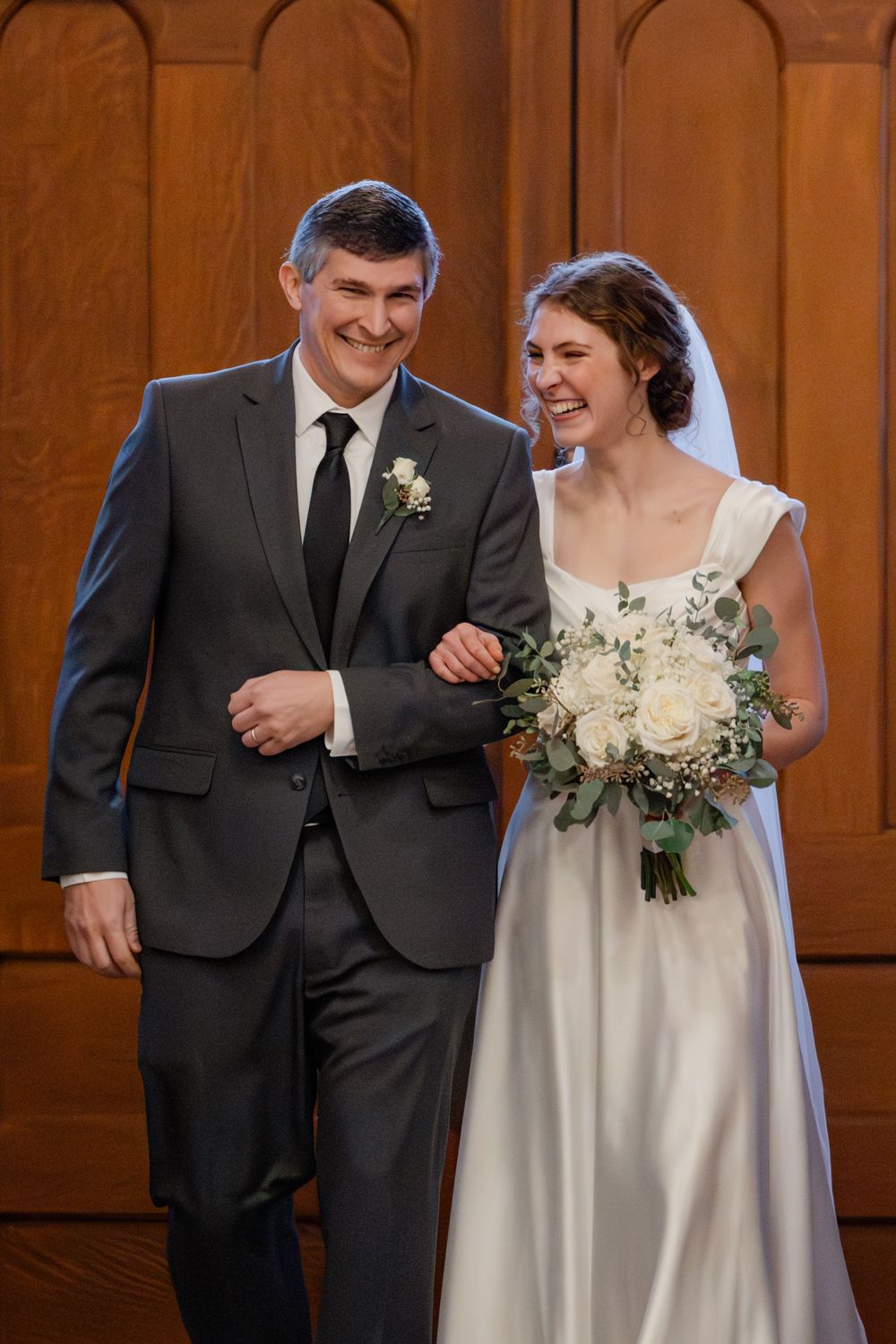 A smiling bride holding white roses walks arm-in-arm with a man in a gray suit at a wedding ceremony.