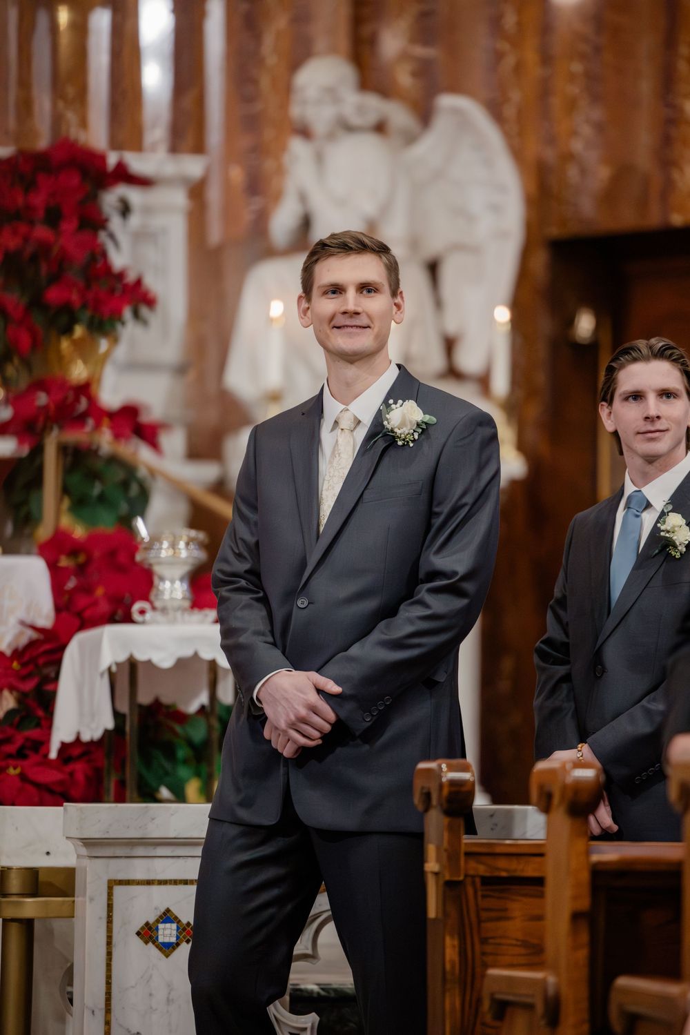 Smiling groom in gray suit with boutonniere stands at church altar during wedding ceremony with groomsman beside him.