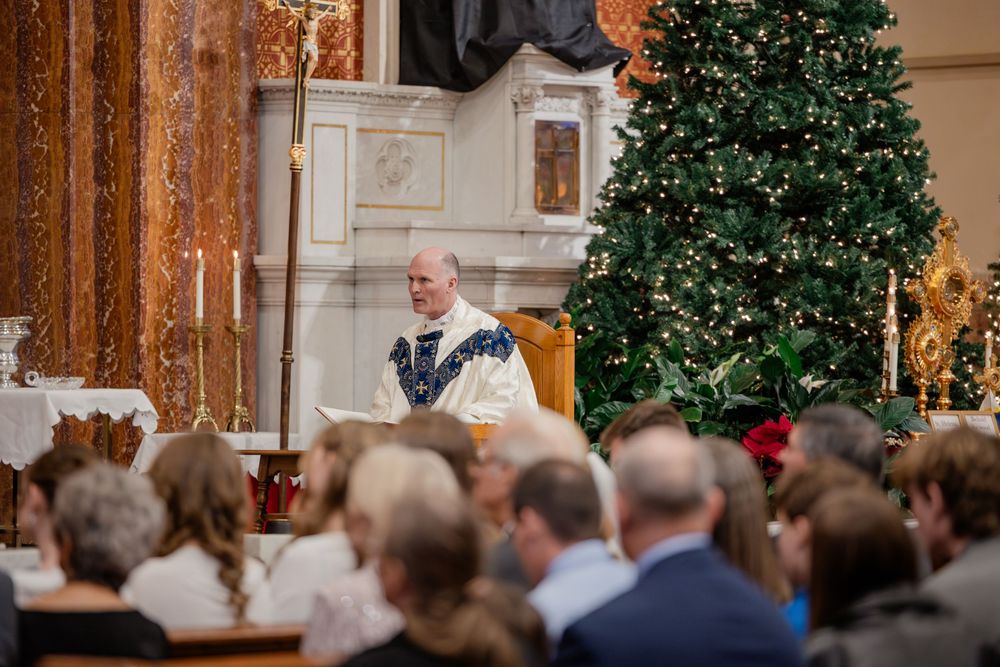 Priest delivers Christmas Mass sermon at decorated church altar with large Christmas tree and congregation.