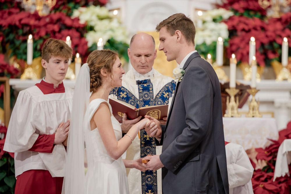 Bride and groom exchange vows at church altar with officiant and altar boy, surrounded by red poinsettias.