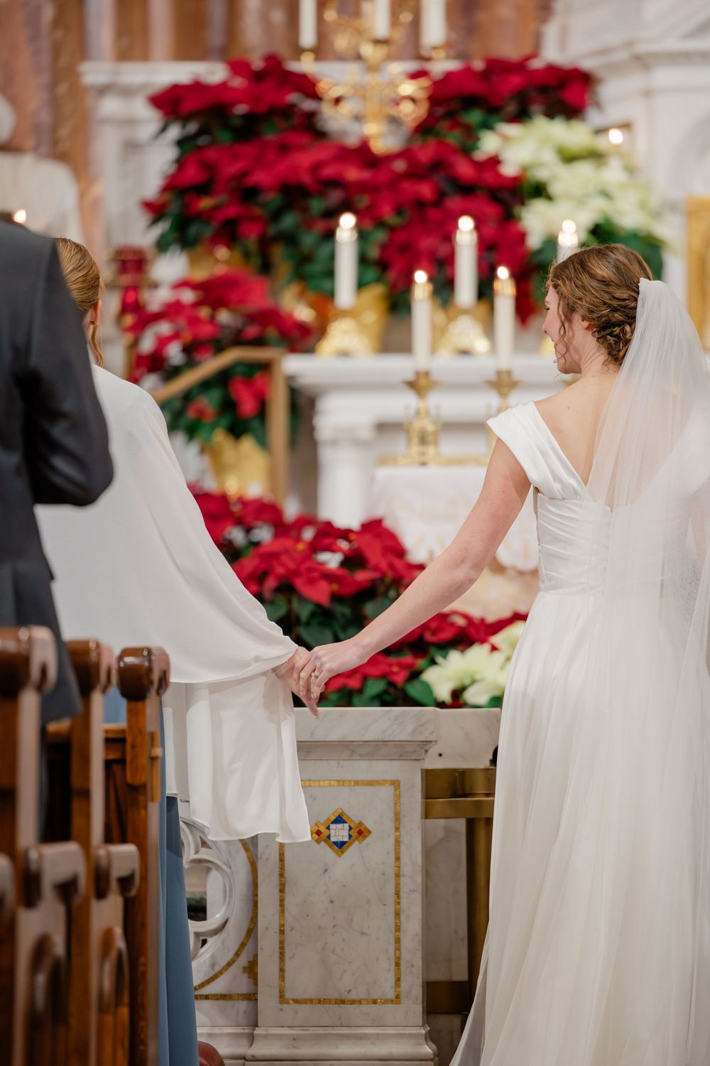 Two brides hold hands at a church altar adorned with red poinsettias and candles during a wedding ceremony.