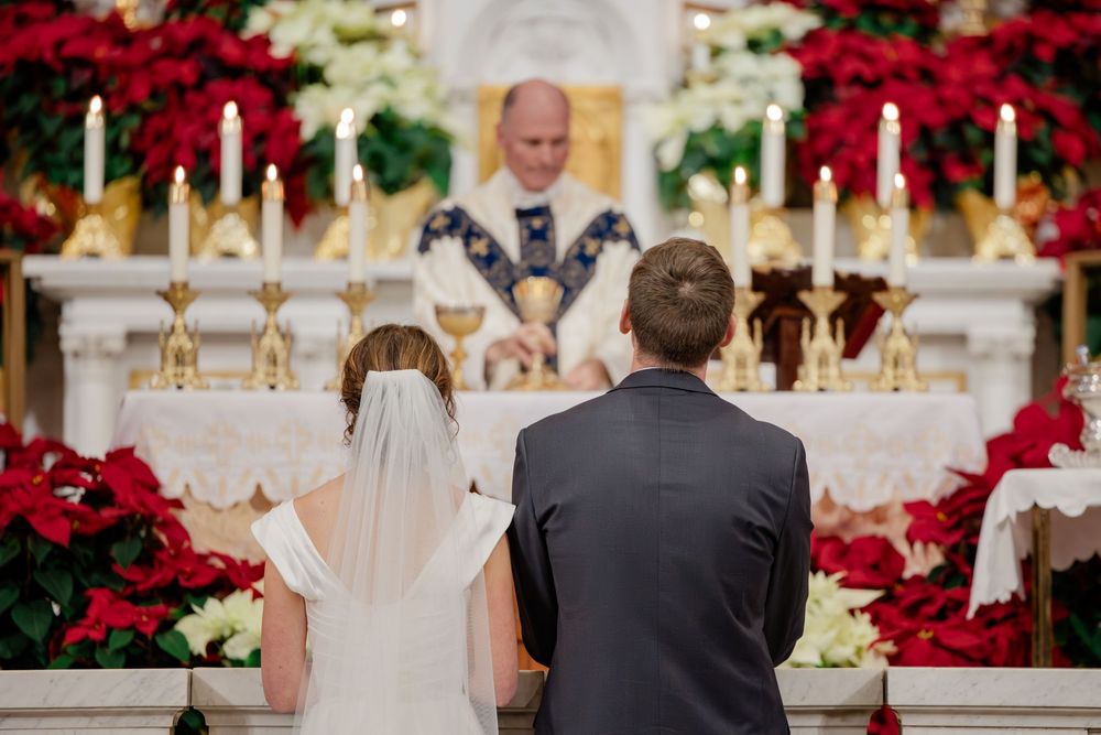 Bride and groom kneeling at altar during Catholic wedding ceremony with poinsettias and candles.