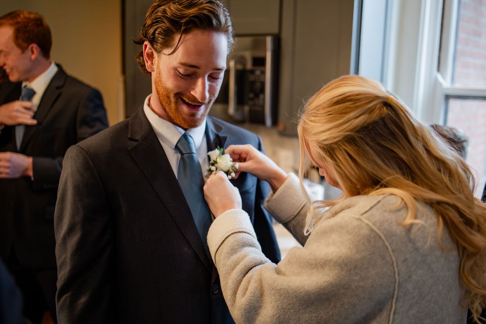 A woman pins a boutonniere on a smiling groom in a dark suit and blue tie during wedding preparations.