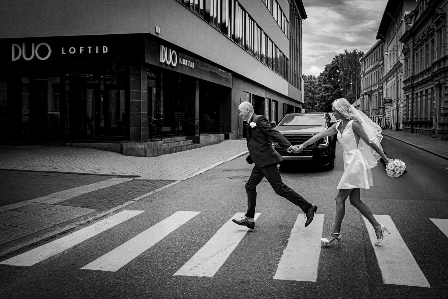 Black and white photo of a couple crossing a zebra crossing, man kicking a soccer ball, woman in white dress.