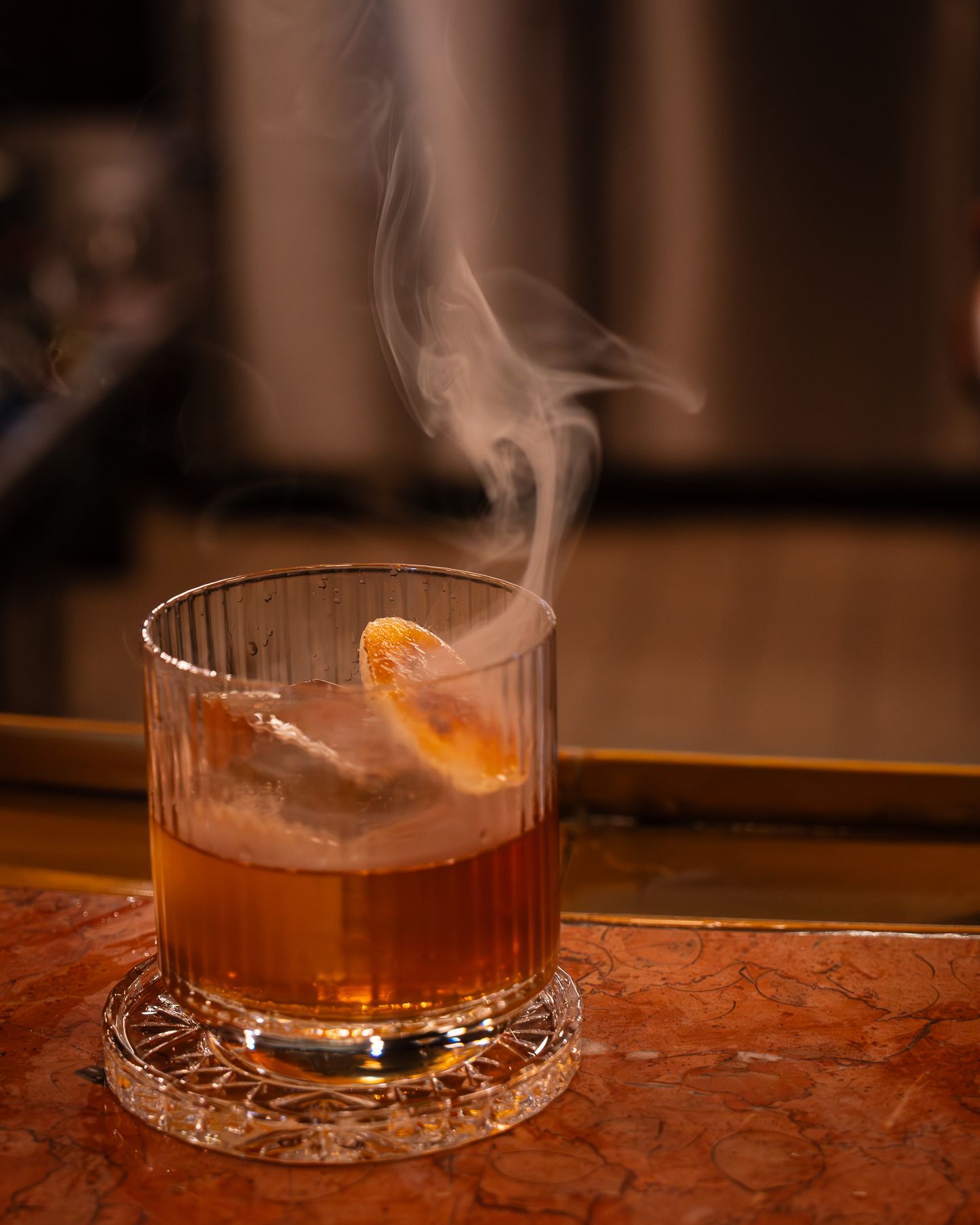 Smoky whiskey cocktail with orange peel garnish in ribbed crystal glass on marble surface.