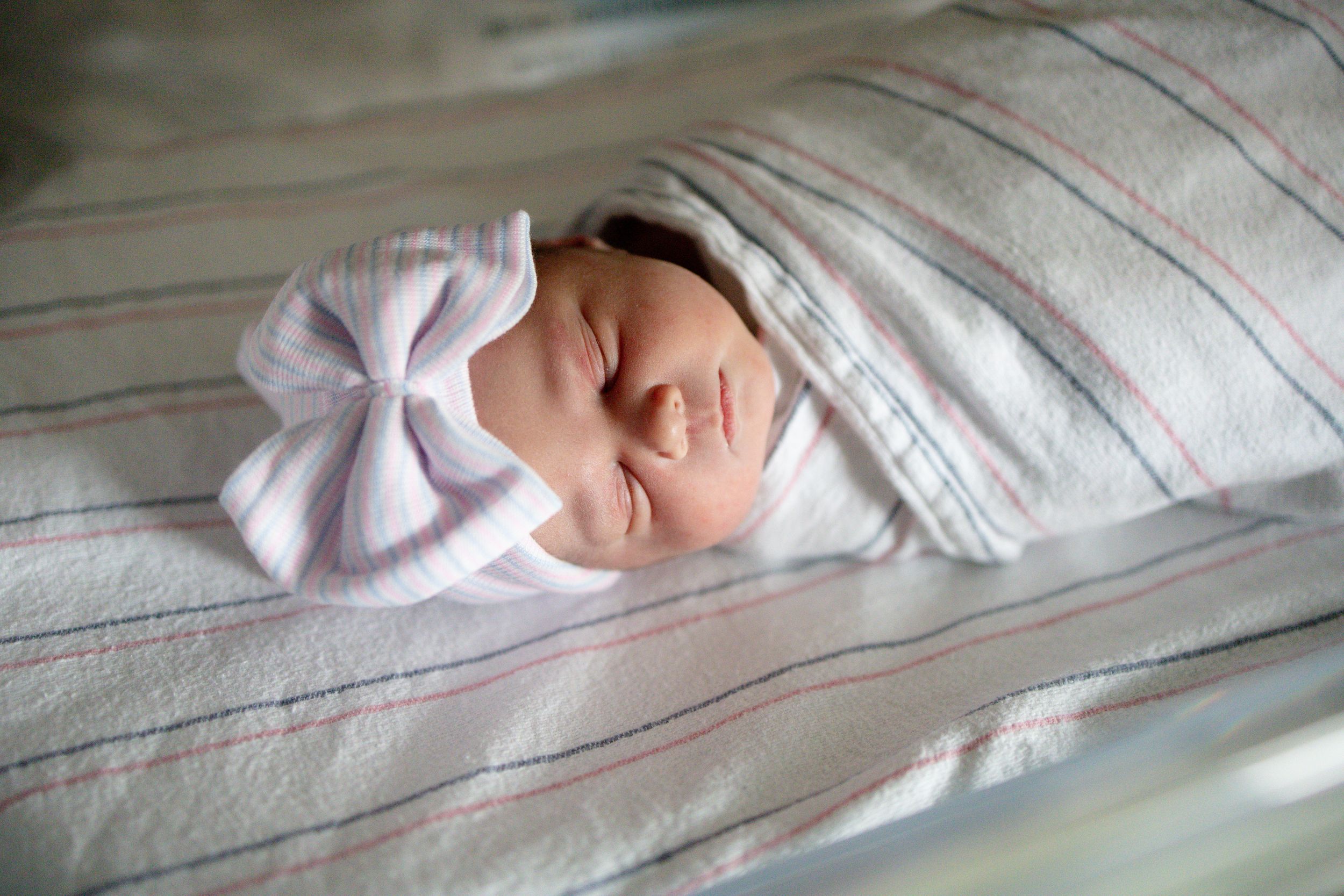 Peaceful newborn baby sleeping swaddled in striped blanket wearing white bow headband in hospital nursery.