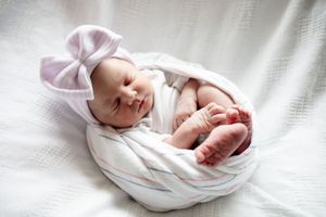 Sleeping newborn baby girl in white swaddle with lavender bow headband showing tiny feet.