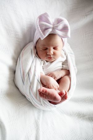 Sleeping newborn baby swaddled in white wrap with pink bow hat on white blanket.