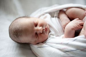 Close-up of sleeping newborn baby partially swaddled in white muslin blanket with tiny fingers visible.