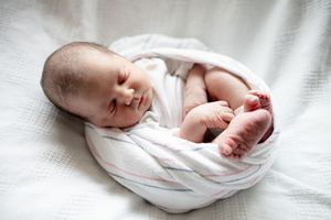 Newborn baby wrapped snugly in white swaddle blanket lying on soft white surface with legs peeking out.