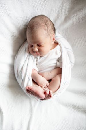 Newborn baby sleeping curled up in white swaddle wrap on soft white textured blanket.