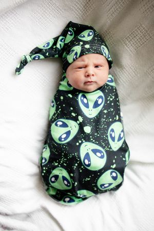 Newborn baby swaddled in black alien print wrap with matching knotted hat on white blanket.