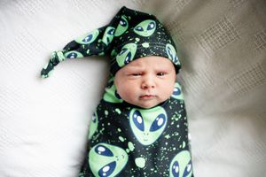 Newborn baby swaddled in black and green alien print wrap with matching knotted hat.