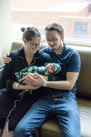 New parents sitting on a couch in a hospital room, lovingly holding their swaddled newborn baby.