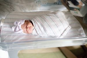 Sleeping newborn baby swaddled in striped hospital blanket with white bow hat in bassinet.