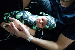 Smiling newborn baby swaddled in alien print blanket being held by father in hospital.