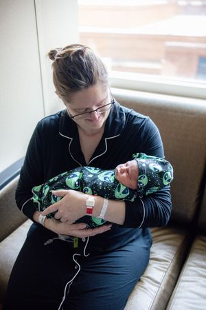 New mother in black pajamas holds swaddled newborn in alien-print blanket while sitting in hospital room.